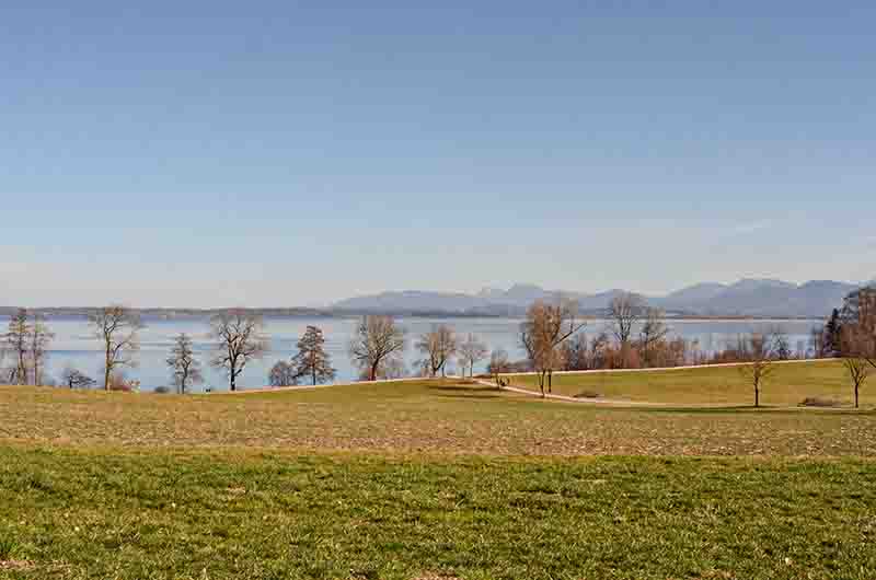 Panoramablick auf den Chiemsee See mit kahlen Bäumen am Ufer, dahinter eine entfernte Alpenkette unter blauem Himmel, davor weite grüne und braune Felder.